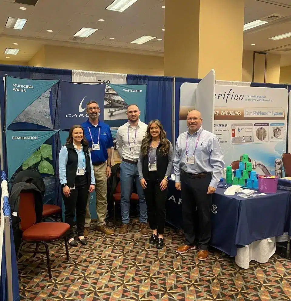 Five people stand smiling in a conference room, posing in front of booths with banners related to water and remediation technologies. They are dressed in business casual attire. The floor is carpeted with a geometric pattern.