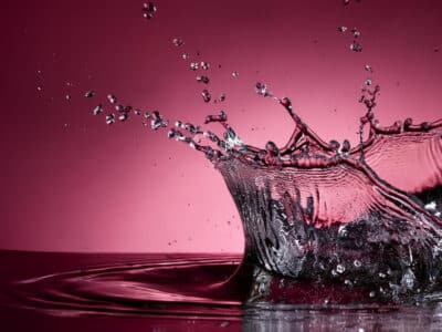 A splash of water captured in mid-air against a deep red background. Water droplets scatter outward, creating dynamic patterns. The surface below reflects the splash, enhancing the image's vibrancy and movement.