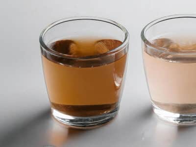 Three clear glasses are lined up on a light gray surface. The first glass contains brown liquid, the second glass has a light pink liquid, and the third glass holds clear liquid, possibly water.