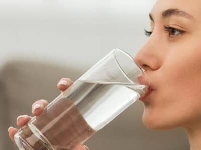 A woman with a ponytail is drinking a glass of water with a blurred background.