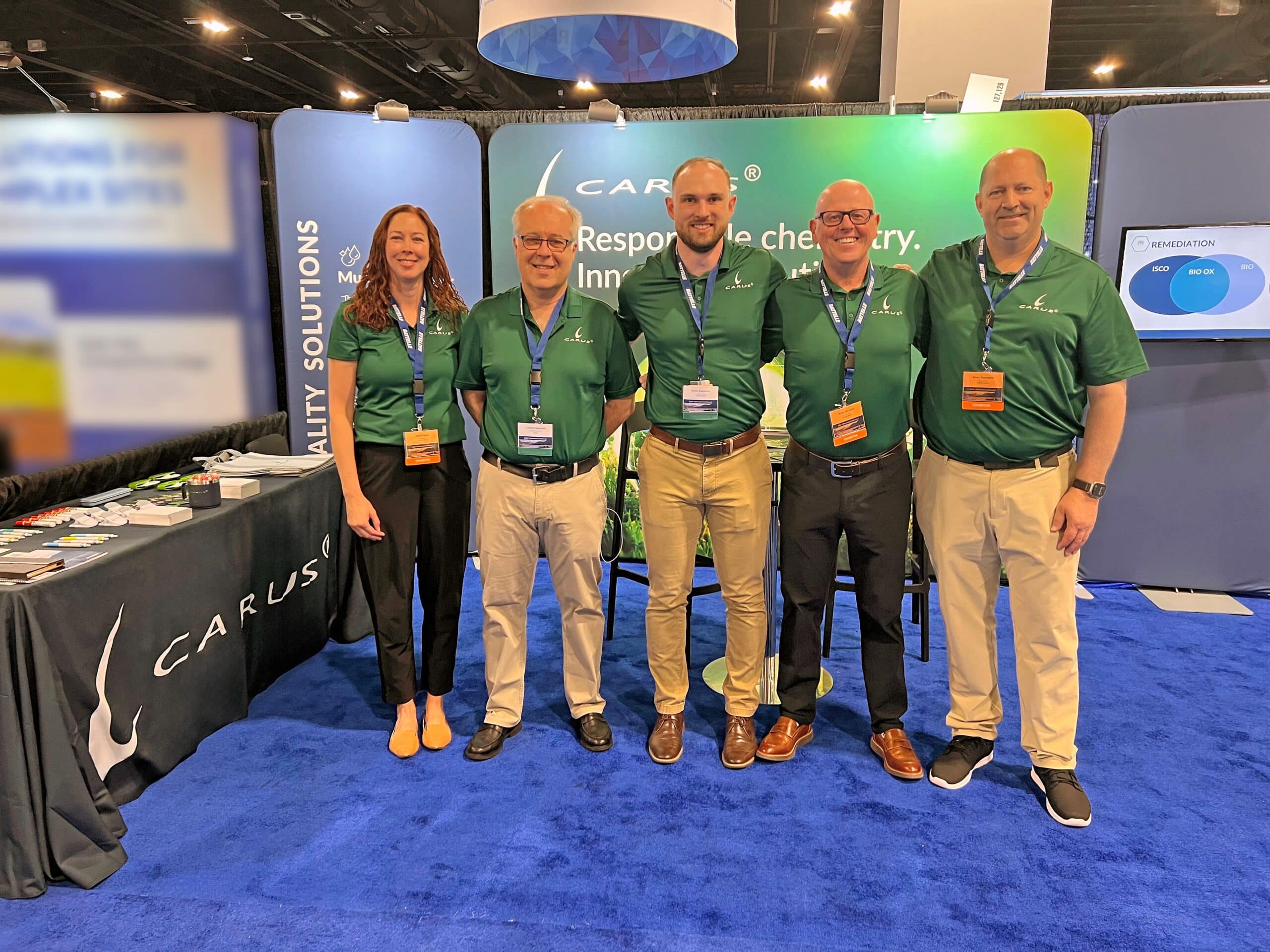 Five people wearing green Carus shirts pose and smile together at a trade show booth with a blue carpet, branded table, and backdrop displaying the Carus logo and slogan.