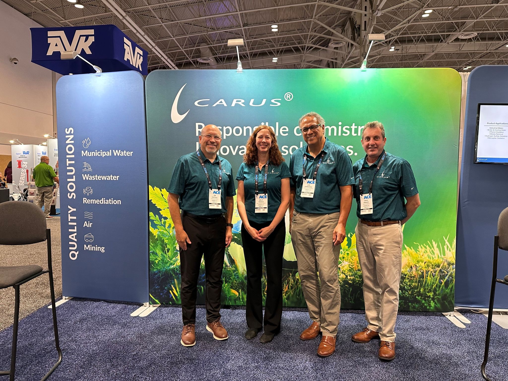 Four people in teal shirts and black pants stand smiling in front of a green and blue Carus company booth display at a convention center. The display lists solutions for water, wastewater, remediation, air, and mining.