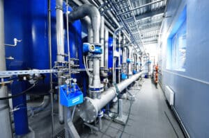 Large industrial water treatment facility with blue tanks, stainless steel pipes, and monitoring equipment in a clean, bright room with tiled floors and a window letting in natural light.