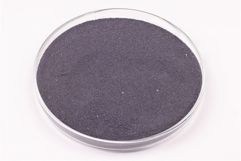 A clear glass petri dish filled with fine, dark gray powder, sitting on a white surface.