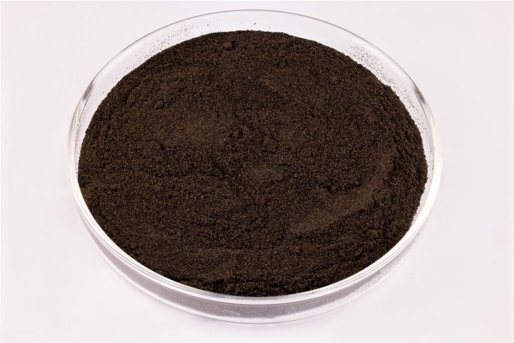 A clear glass petri dish filled with fine, dark brown or black powder sits on a white background.