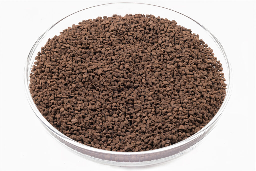 A clear glass dish filled with small, dark brown granules, resembling instant coffee granules, placed on a white background.