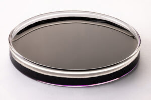 A glass petri dish filled with a dark, almost black liquid sits on a white surface. The liquid appears smooth and reflective, with a faint purple tint around the edges.