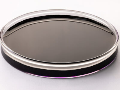 A glass petri dish filled with a dark, almost black liquid sits on a white surface. The liquid appears smooth and reflective, with a faint purple tint around the edges.