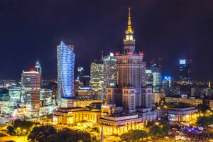 A night view of Warsaw’s skyline with illuminated modern skyscrapers and the historic Palace of Culture and Science prominently lit in the center.