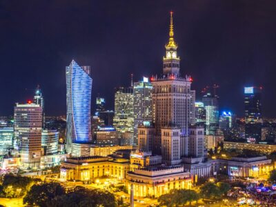 A night view of Warsaw’s skyline with illuminated modern skyscrapers and the historic Palace of Culture and Science prominently lit in the center.