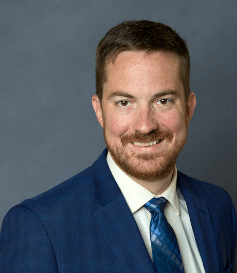 A man with short brown hair and a beard, wearing a blue plaid suit, white shirt, and blue patterned tie, smiles at the camera against a plain gray background.