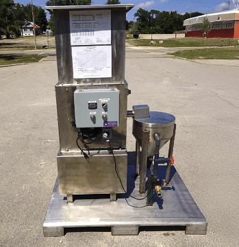A stainless steel industrial machine with control panel and attached cylindrical component stands outdoors on a pallet, with grass, trees, and buildings visible in the background.