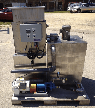 Industrial equipment with control panel, wiring, and pipes mounted on a metal platform outdoors. There is a blue motor, silver tank, and other mechanical components. The background shows cars, buildings, and a dirt area.
